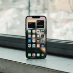 iPhone 16e hiển thị màn hình chính trước cửa sổ, thể hiện giao diện người dùng và thiết kế mặt trước.