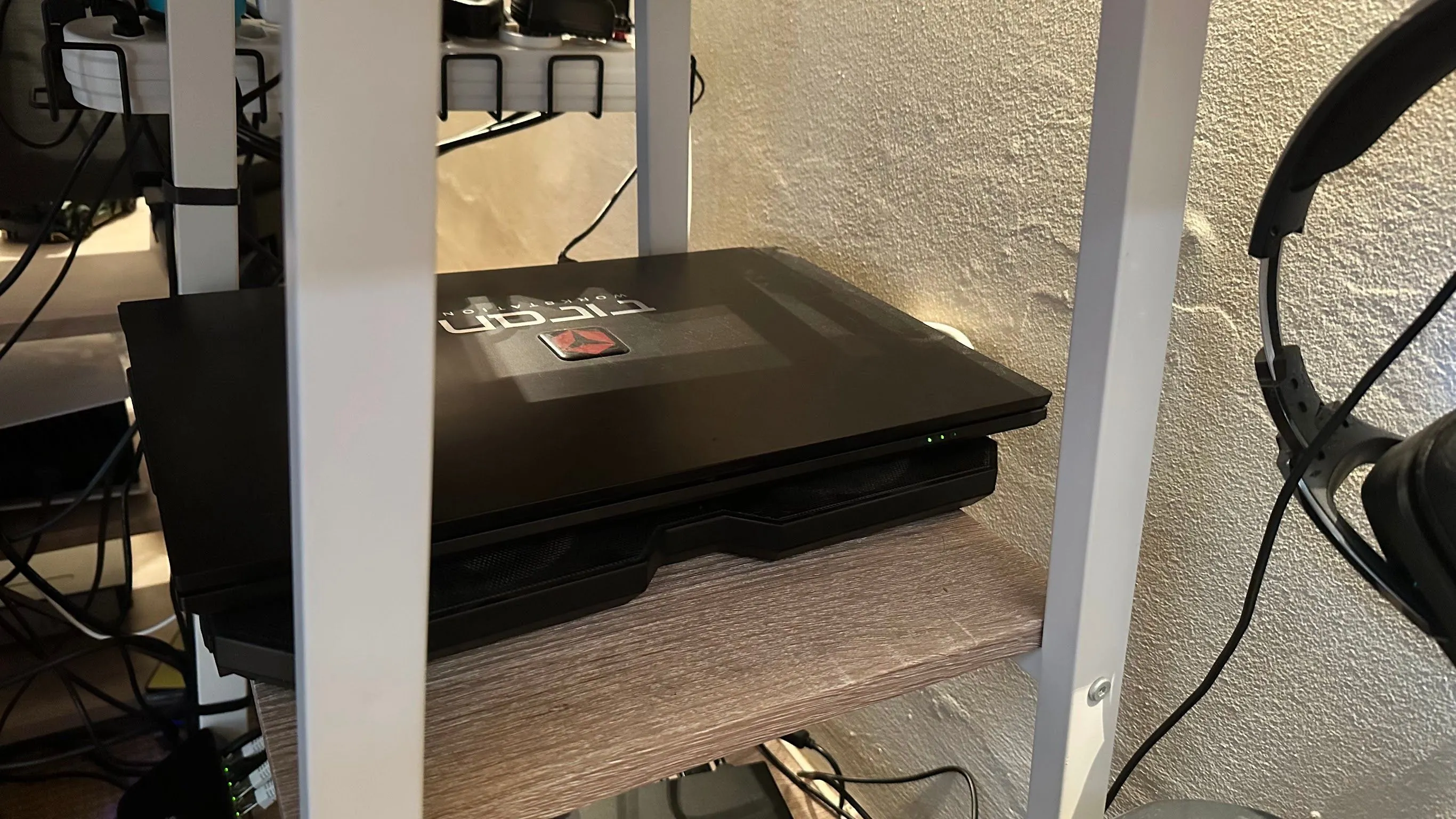 Laptop gập nắp đặt gọn gàng trên đế tản nhiệt, giấu trong kệ bàn làm việc để tối ưu không gian.